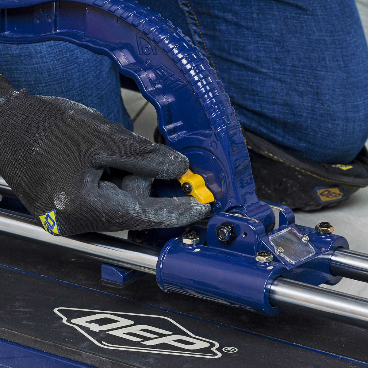 Installer making an adjustment to the height of the cutting wheel on the QEP 28" Big Clinker Tile Cutter while wearing QEP SureGrip Tiler's Gloves and ProMAX Gel Knee Pads