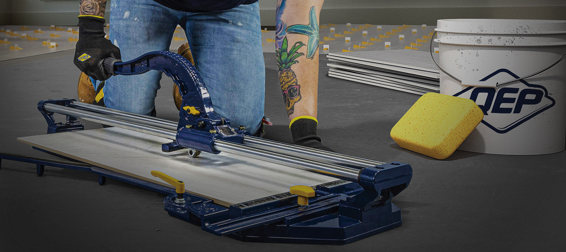QEP tile cutter in use cutting tile shown with QEP QEP SureGrip Tiler’s Gloves, grout sponge, heavy duty plastic mixing bucket, and LASH tile leveling system